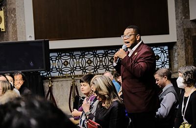 A youn Black man using a microphone speaking to an audience.